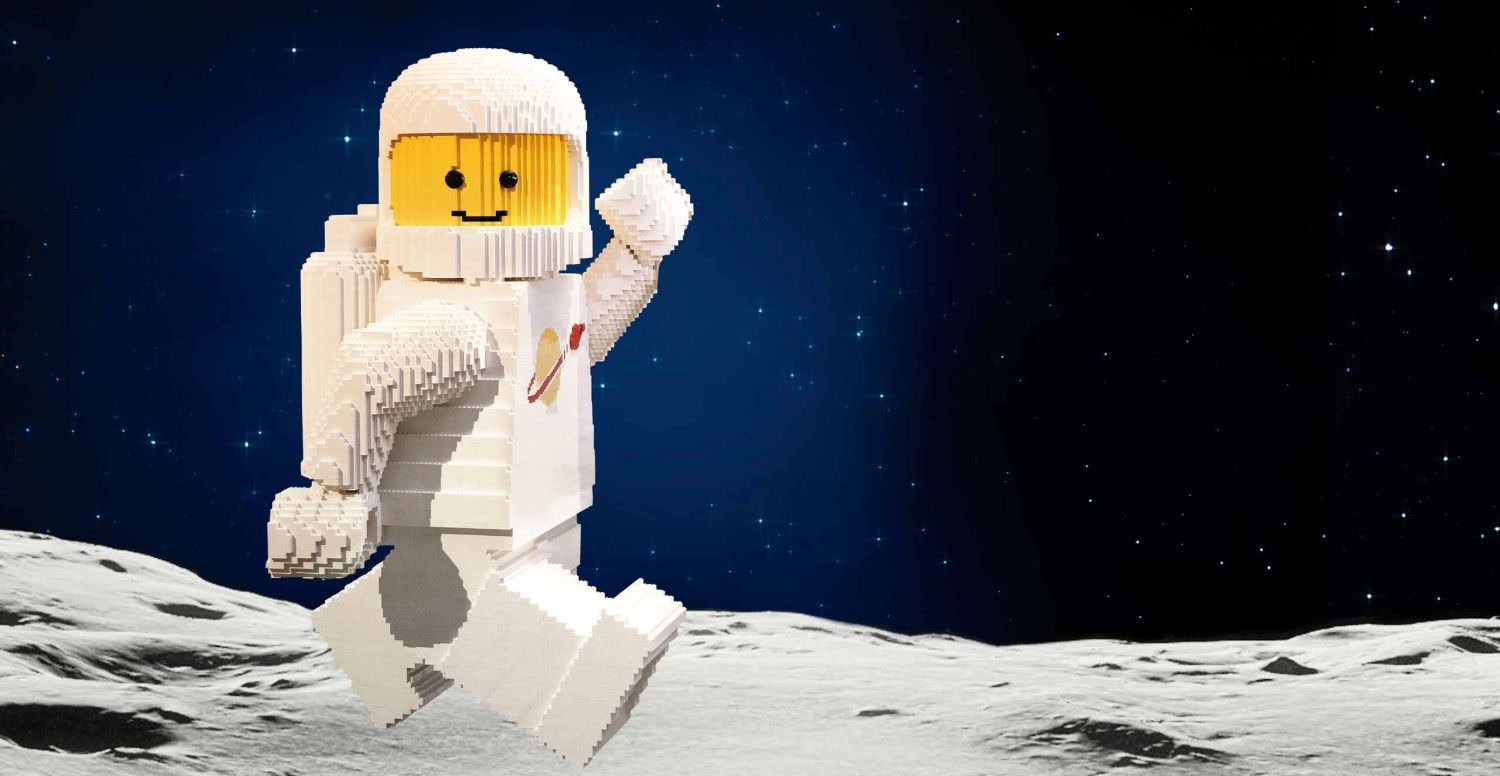 Un grand astronaute Lego blanc qui marche sur un sol lunaire, sous un ciel étoilé.